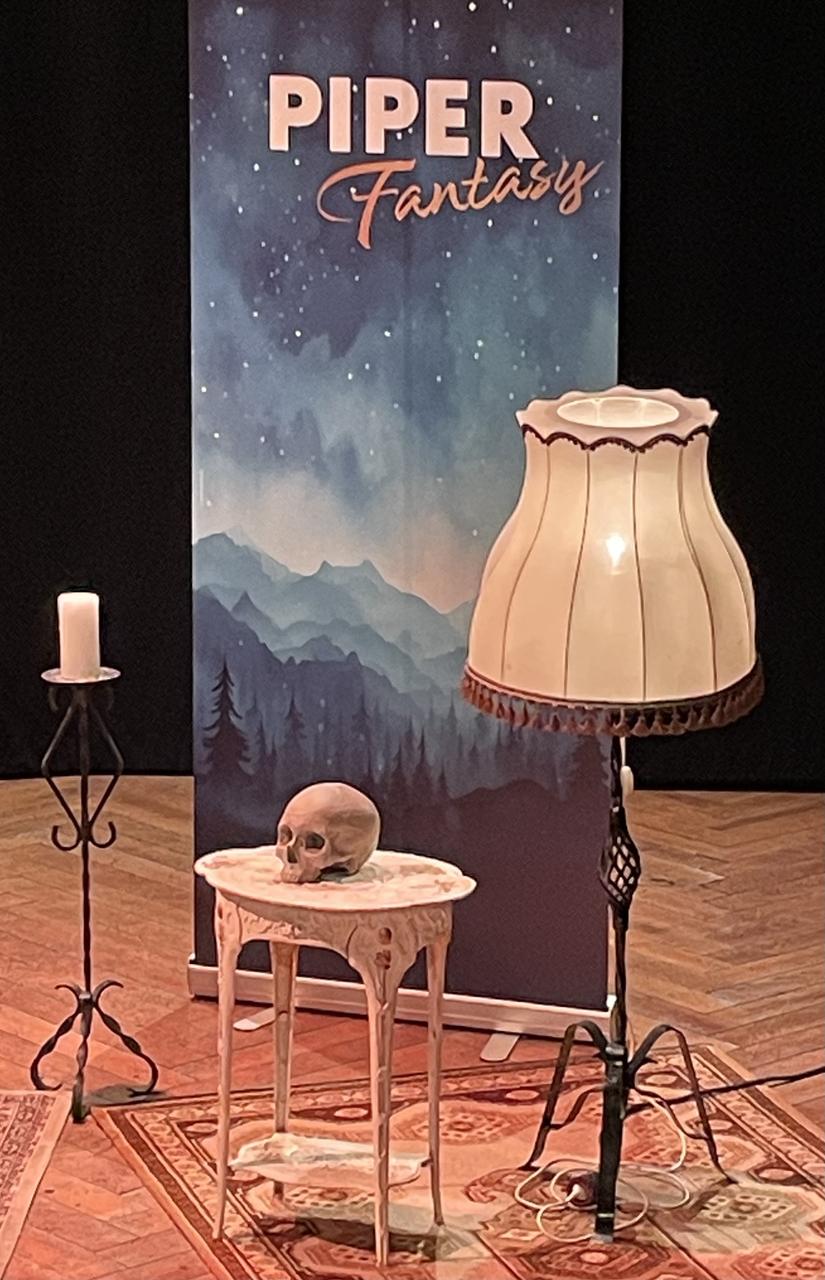 Foto von einem Plakat der PIPER Fantasy Nacht mit dekorativen Elementen davor, wie einer Stehlampe, einem Kerzenständer und ein Tisch mit einem Totenkopf darauf.
